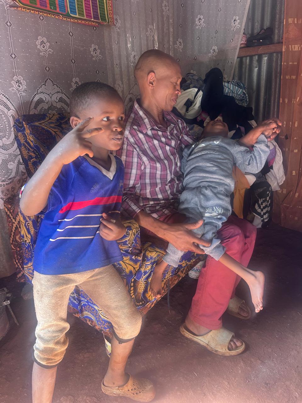 A child with a disability held on an adult’s lap at home in Ruiru, with another child beside them